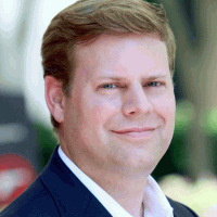 Man with fair skin, smiling slightly, wearing a dark blue blazer and white shirt.