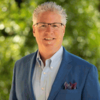 Man with gray hair, glasses, in blue jacket, smiling outdoors.