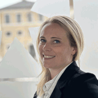 Woman with blonde hair smiles, wearing a suit jacket, near a window with a blurred cityscape.