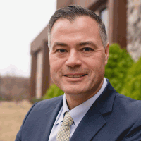 Man in a navy suit, smiling. Outdoors in front of a stone building with green bushes.
