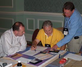 Three men at a table, reviewing documents. One points, another writes, and a third looks on. Conference setting.