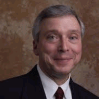 Smiling man in a suit, dark hair, fair skin, against a blurred brown background.