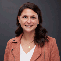 Woman with dark hair smiles, wearing a rust-colored jacket and white shirt, against a gray background.