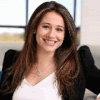 Woman with long brown hair, smiling, wearing a black blazer over a white top.
