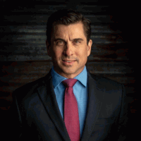 Man in a suit with blue shirt and burgundy tie, looking at the viewer.