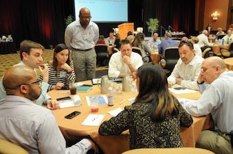 People at a conference sitting at round tables. A speaker stands in the background, looking at the audience.