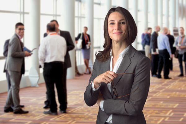 Woman in a business suit smiles, holding glasses, standing in front of a blurred conference setting.
