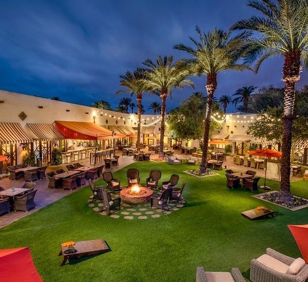 Outdoor restaurant courtyard with palm trees, tables, a fire pit, and evening lighting.