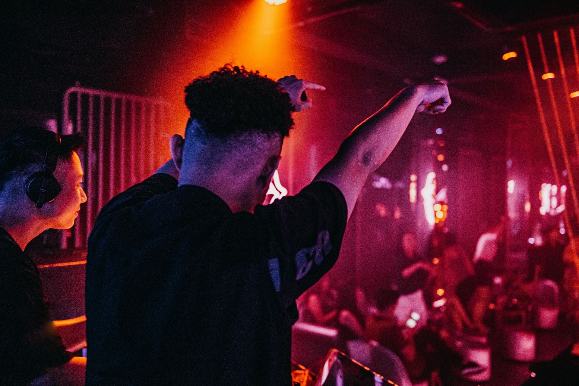 Man in a club with raised arm, pointing. DJ in the background, red and orange lighting, crowd visible.
