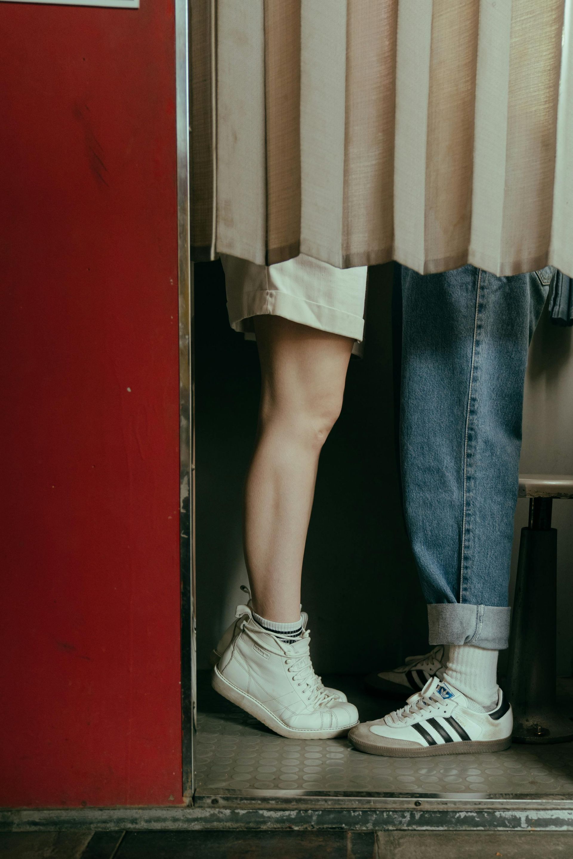 Two people's legs and feet close together in a photo booth; one in shorts and white sneakers, the other in jeans and Adidas sneakers.