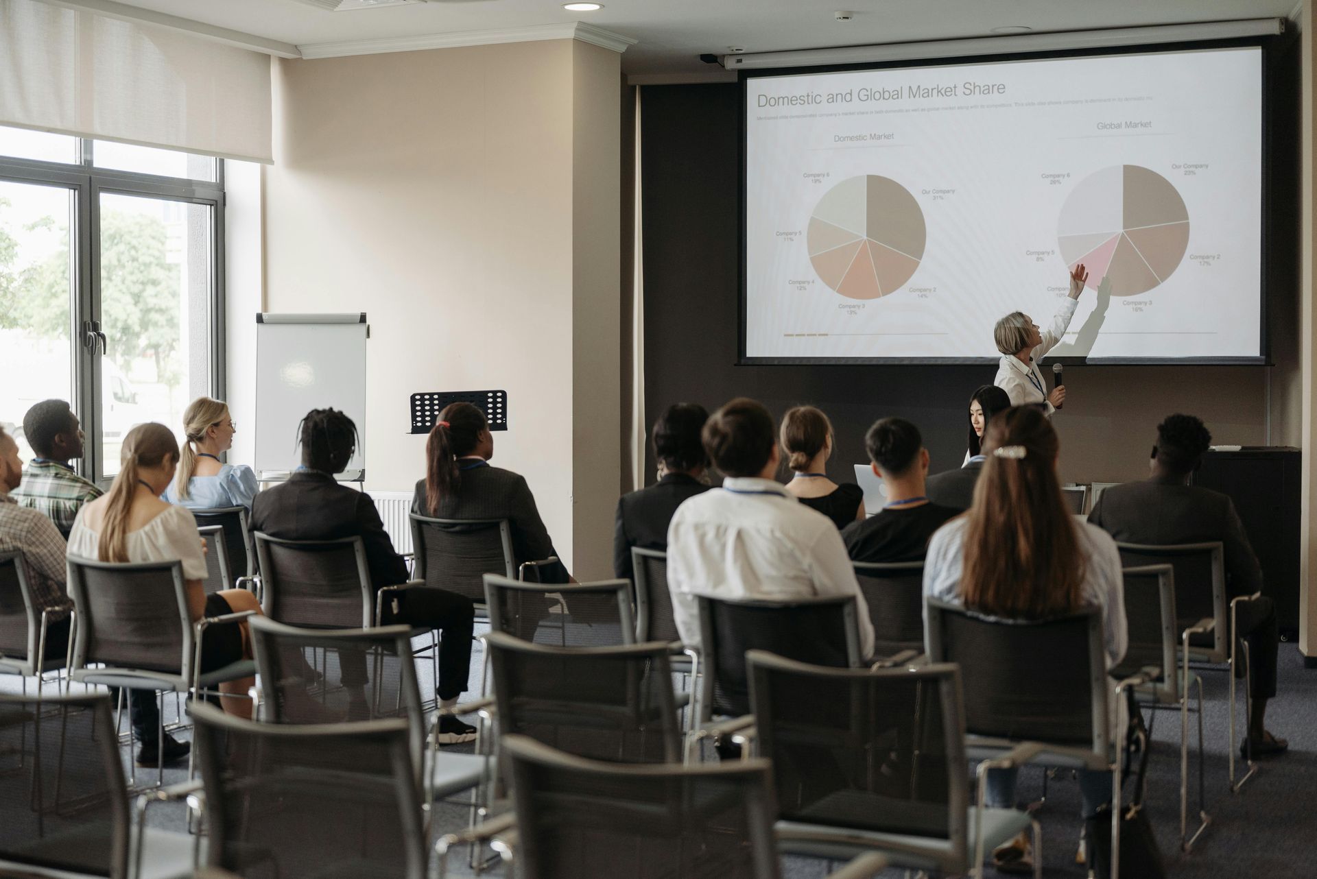 A person presenting data from pie charts on a projector screen to a seated audience in a conference room.
