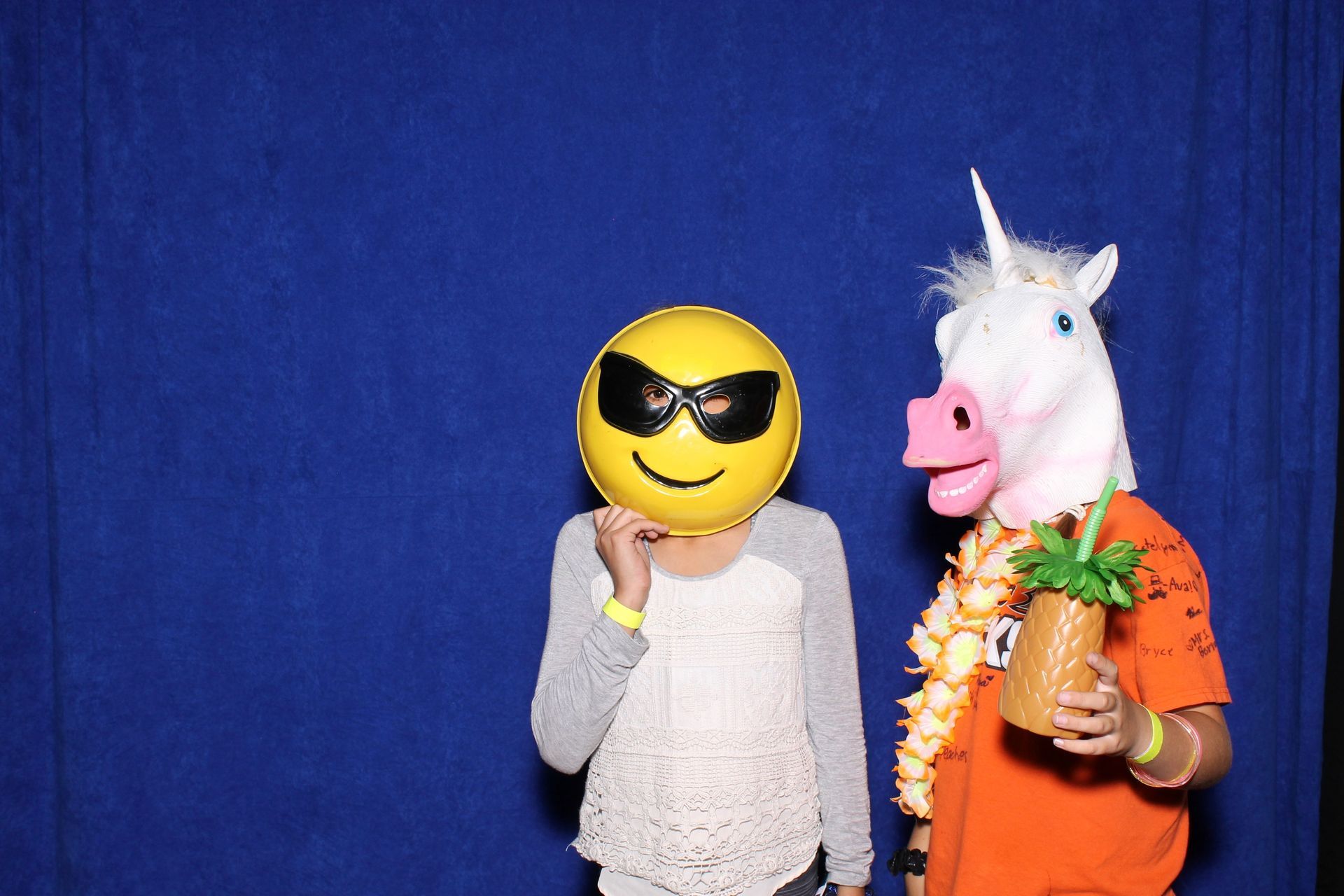Person in emoji mask and person in unicorn mask pose in front of a blue backdrop, holding a cocktail.