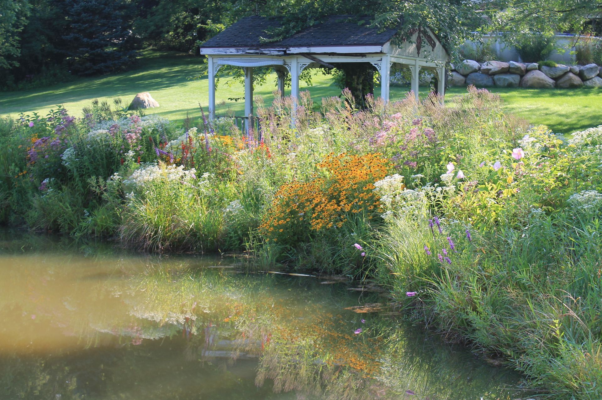 Lush garden with wildflowers bordering a pond, gazebo in the background. Reflective water, sunny setting.
