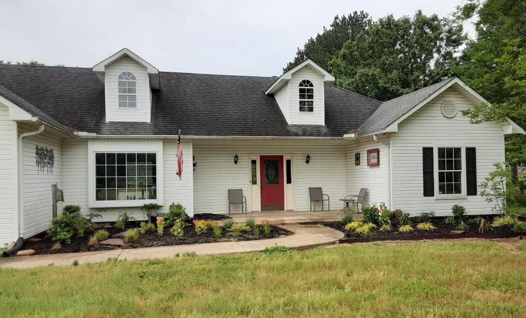 A white house with a red door and black shutters is sitting on top of a lush green field.