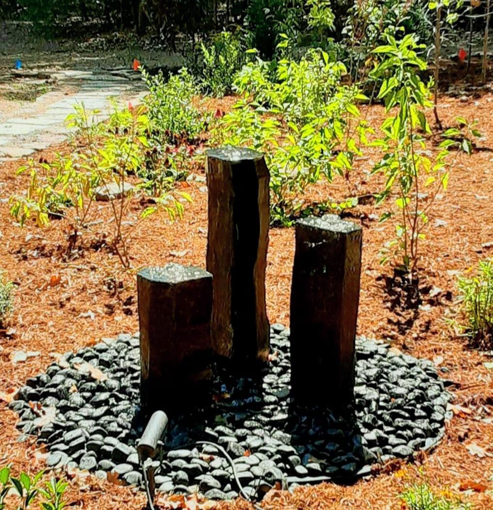 Three dark stone pillars in a garden bed with black pebbles, water feature.