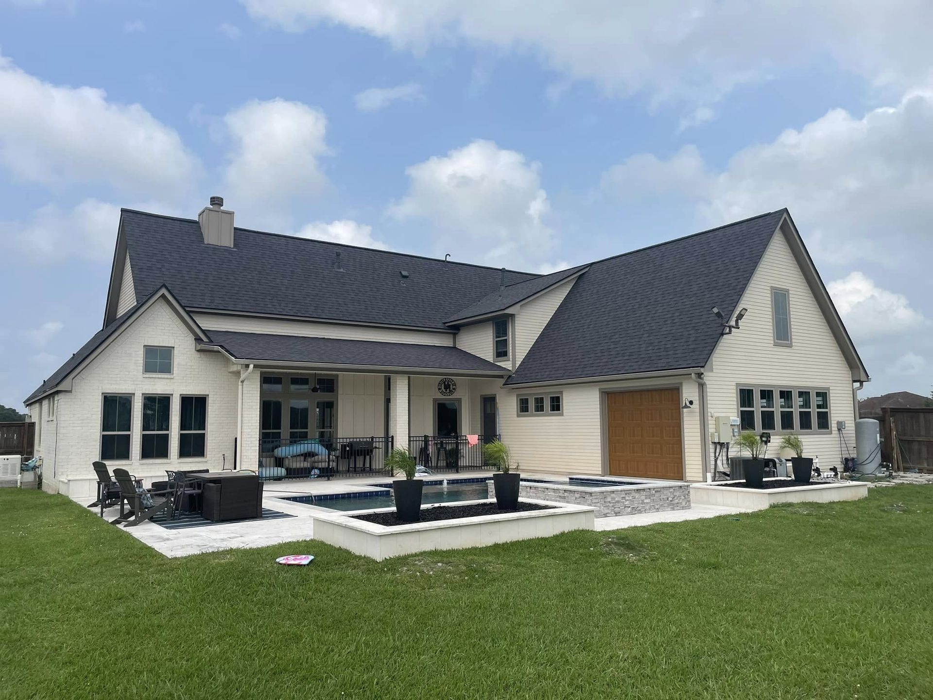 Backyard view of a two-story beige house with black roof, swimming pool, and green lawn under a cloudy sky.