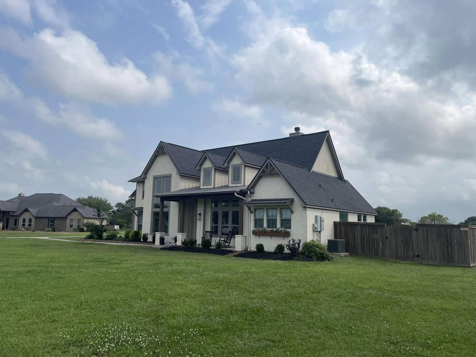 A two-story house with black roof and white exterior sits on a grassy lawn under a cloudy sky.