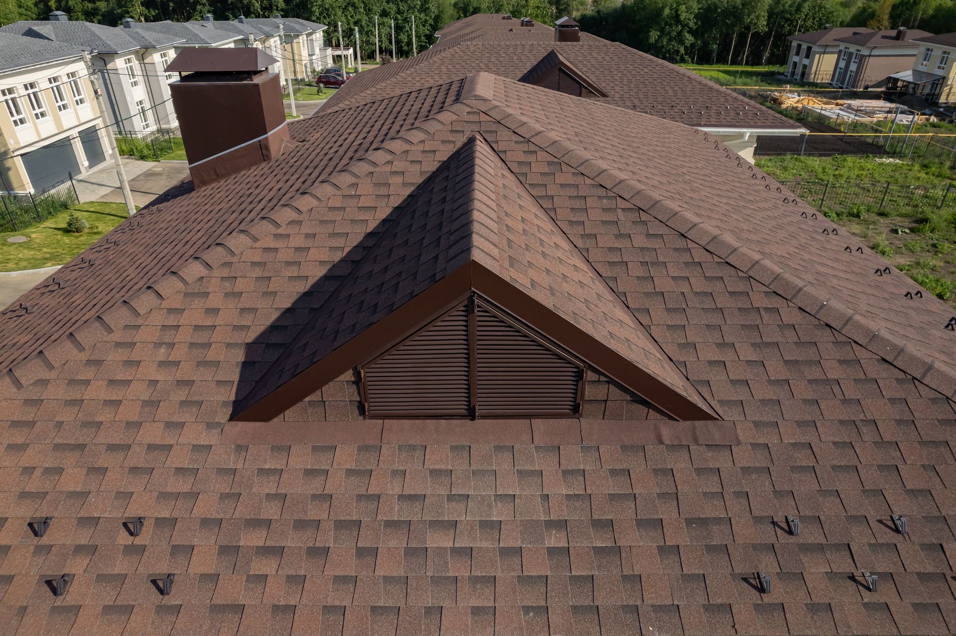 Brown shingled roof with a triangle-shaped window and chimney, overlooking a residential area.