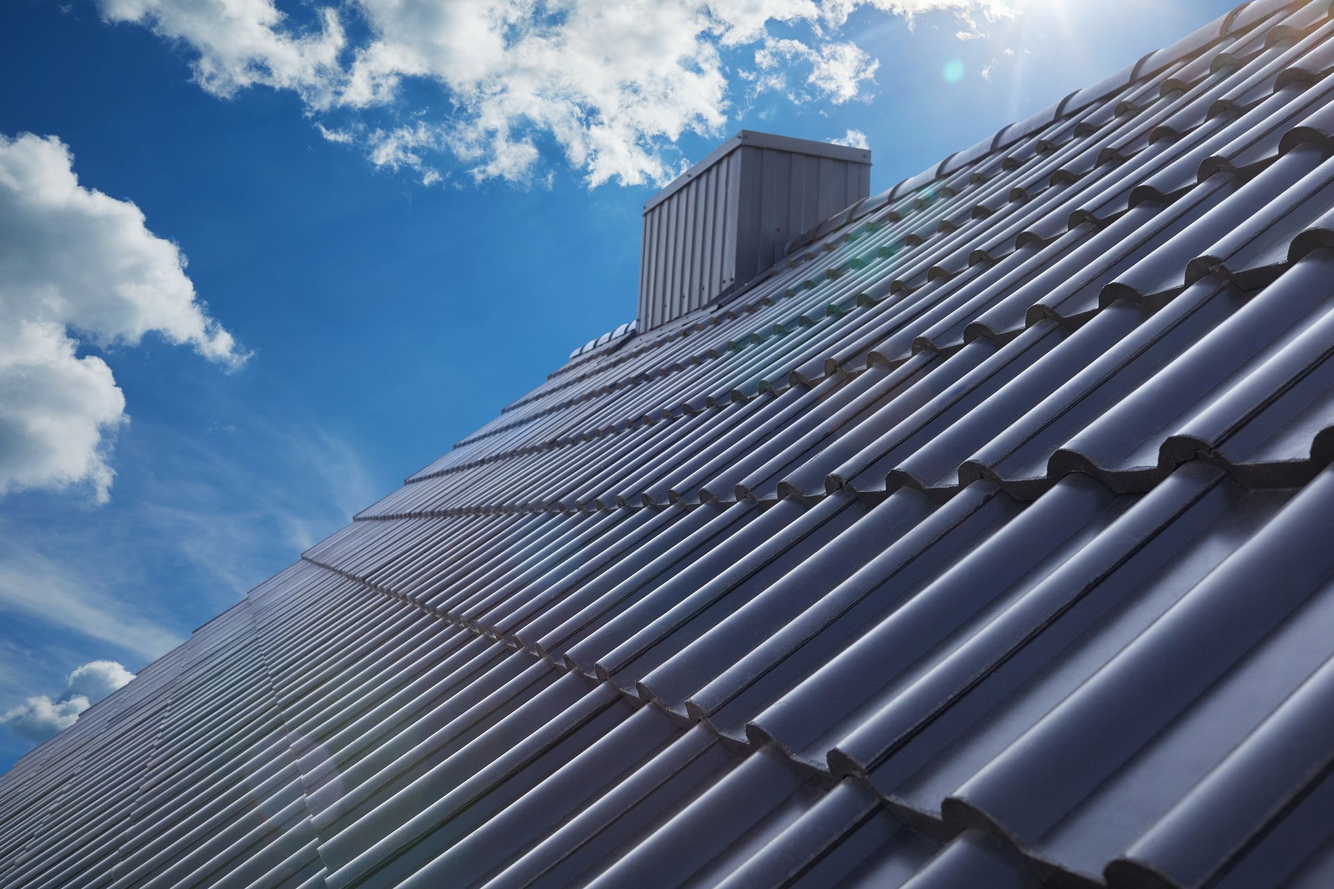 Gray tiled roof with chimney against a blue sky with sunlight.