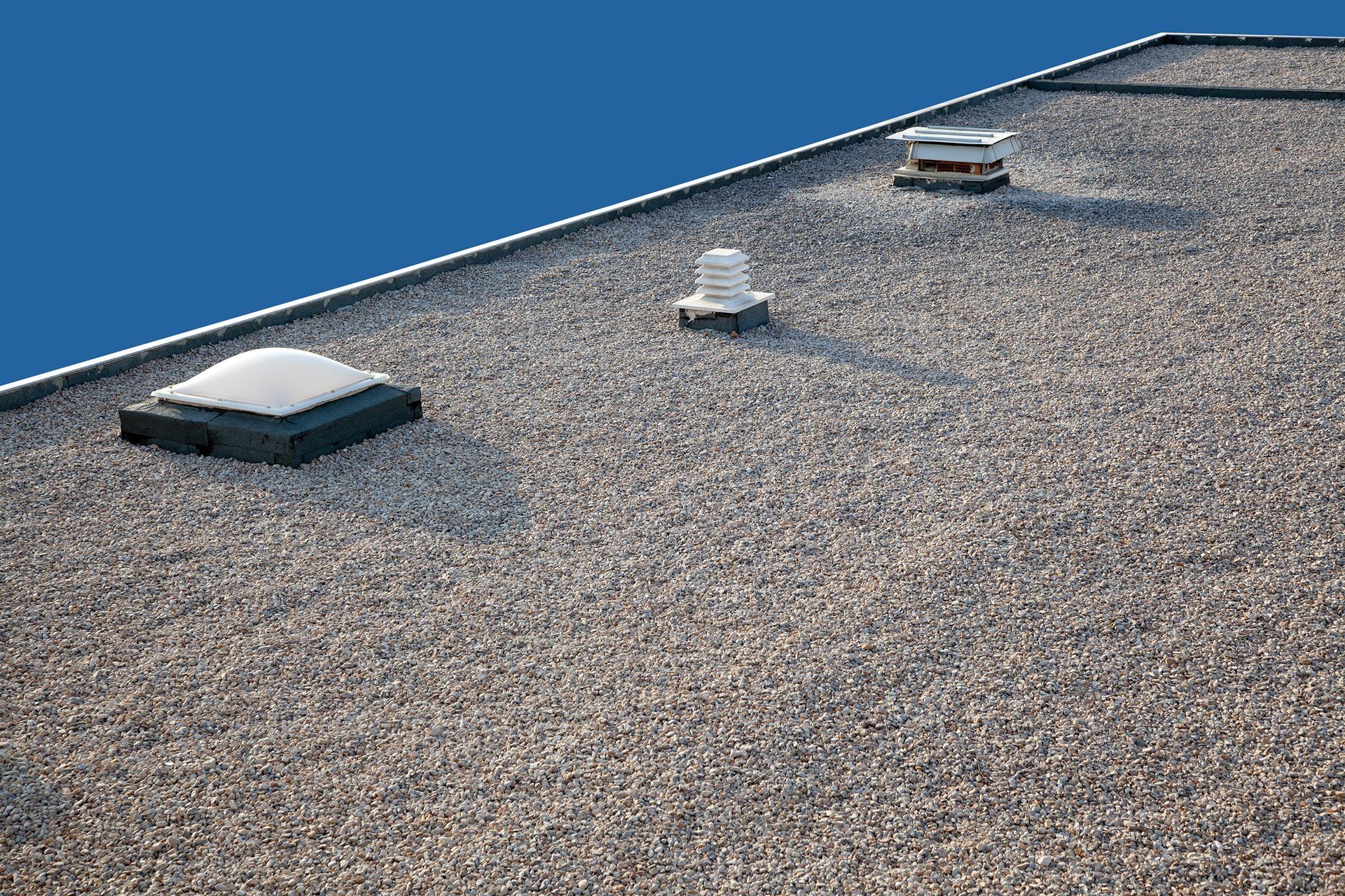 Flat gravel roof with three vents and a skylight against a blue sky.