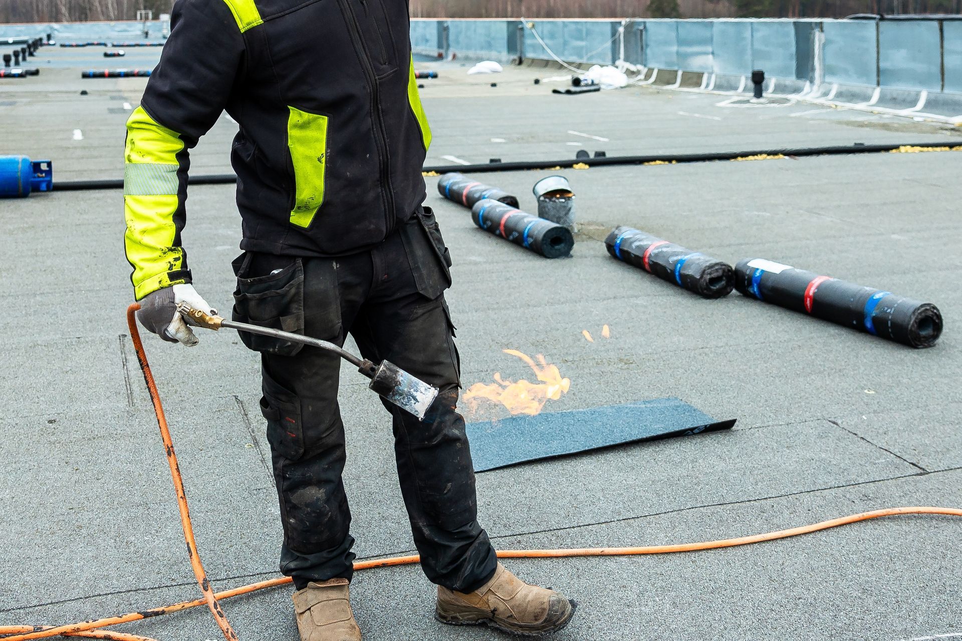 Roofer using a torch to work on a flat roof. They wear black work clothes and yellow safety accents.