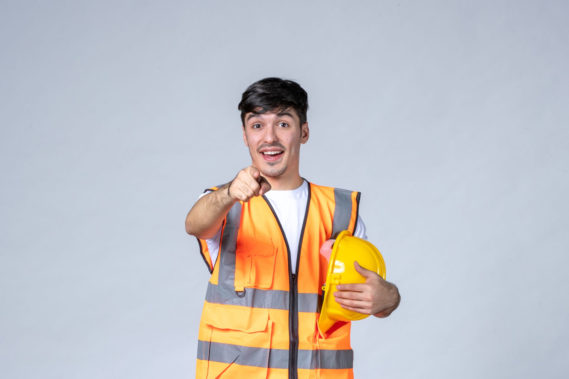 Man in orange safety vest points at viewer, holding a yellow hard hat.