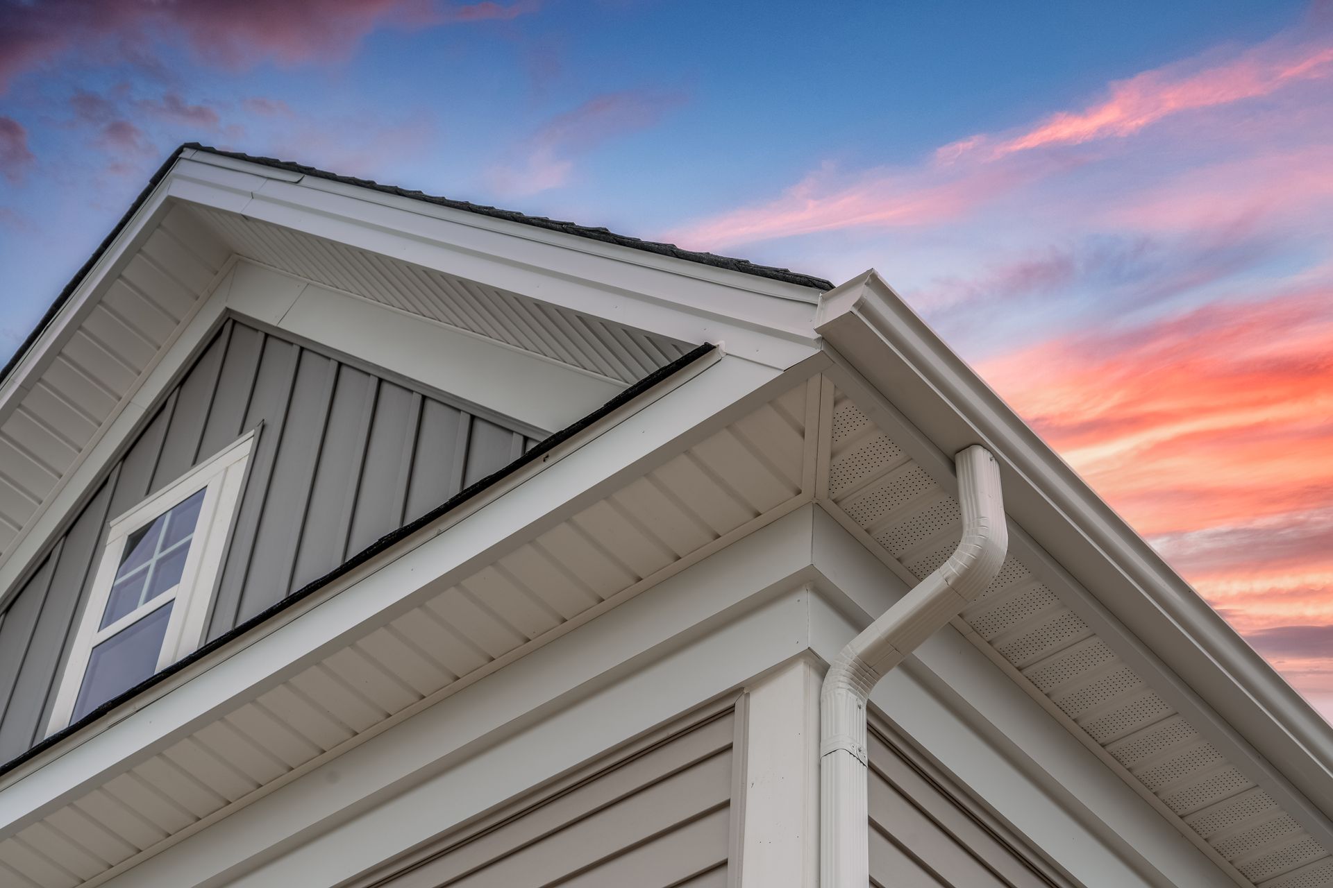Close-up of a house corner with white trim, gray siding, and a gutter, against a sunset sky.