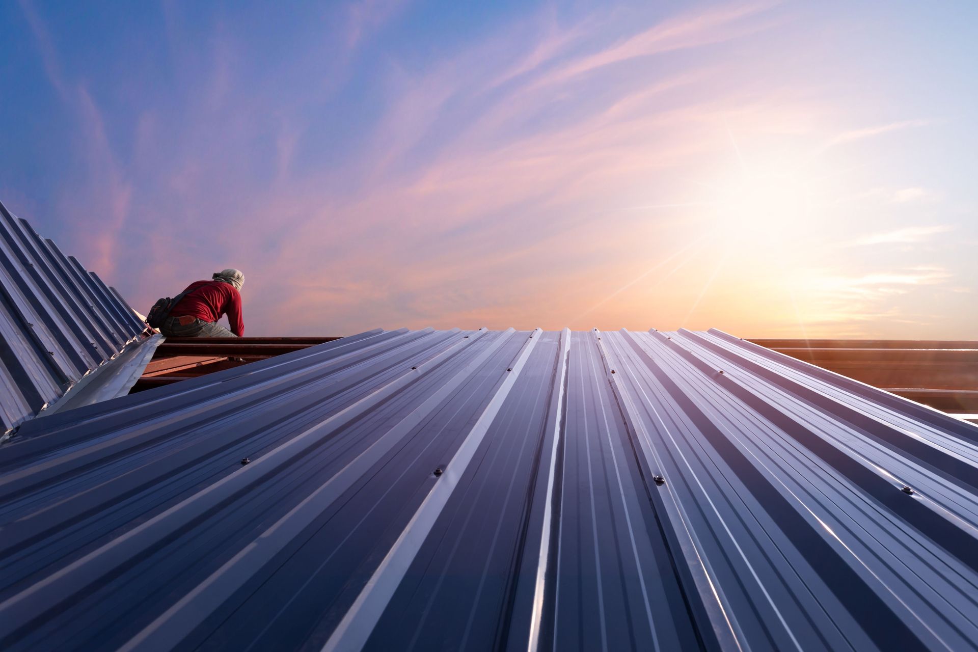 Roofer in red shirt working on a metal roof against a sunset sky.