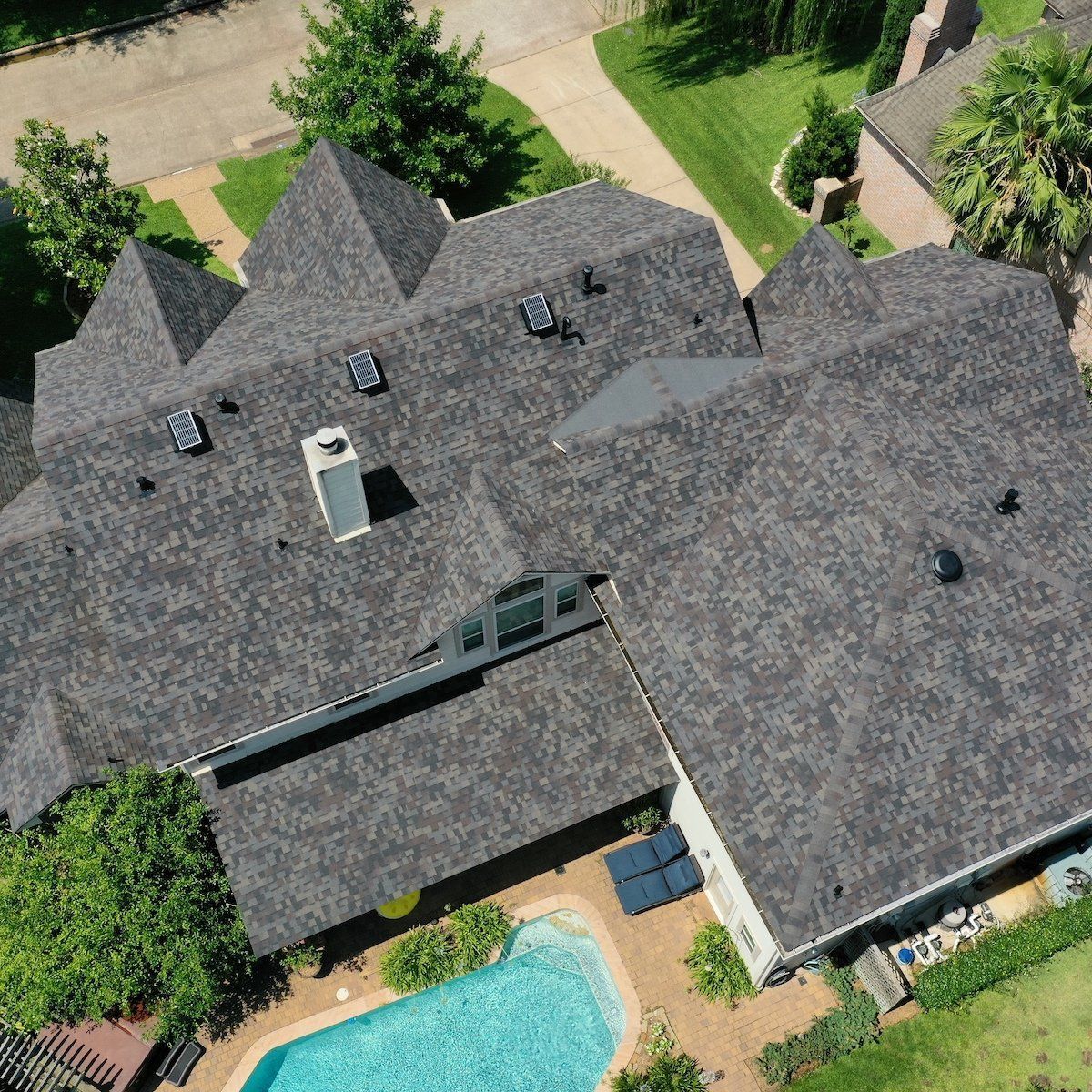 Aerial view of a dark gray asphalt shingled roof on a large house with a pool.