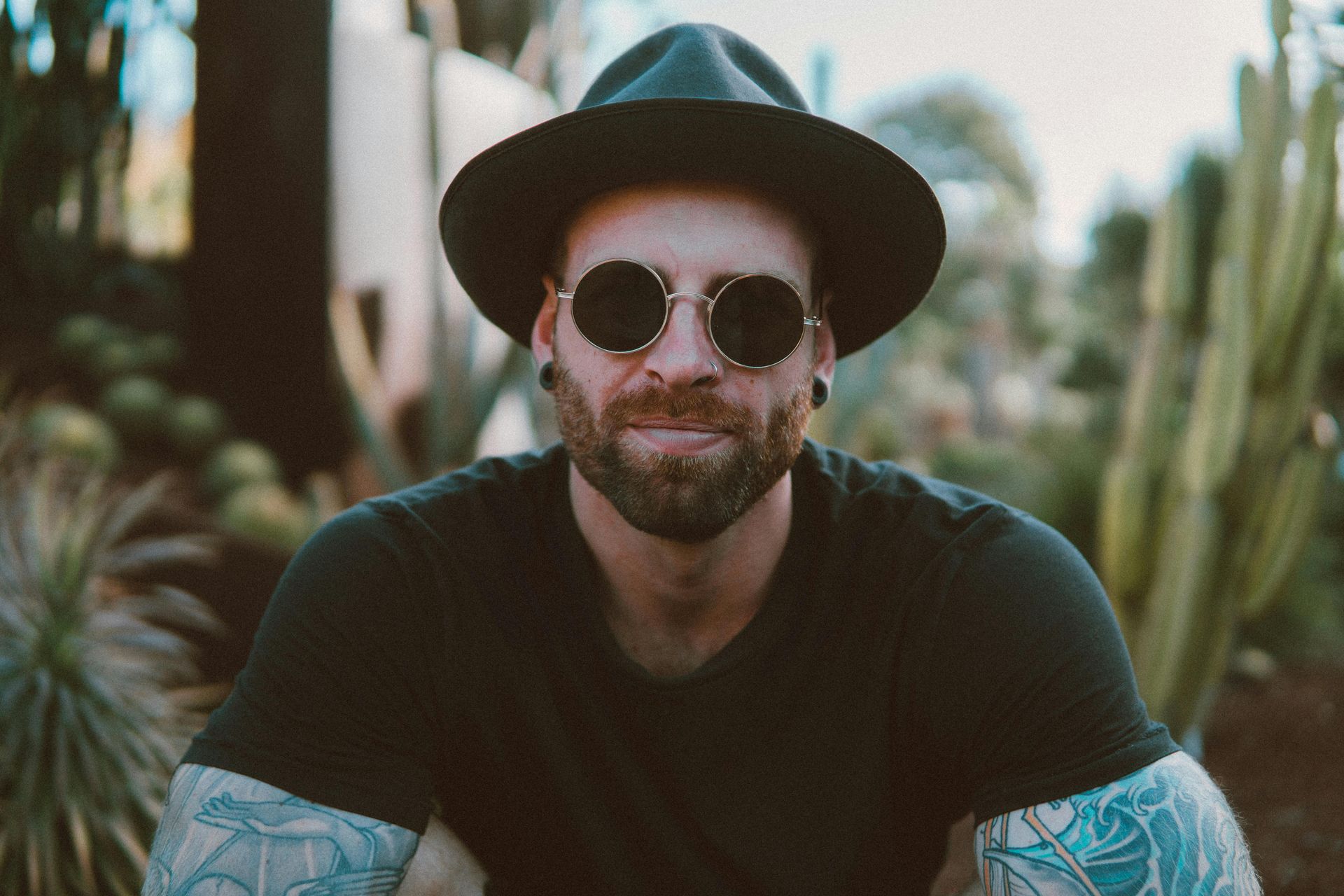 Man with a beard wearing a fedora and sunglasses smiles. Tattooed arms, outdoor setting.