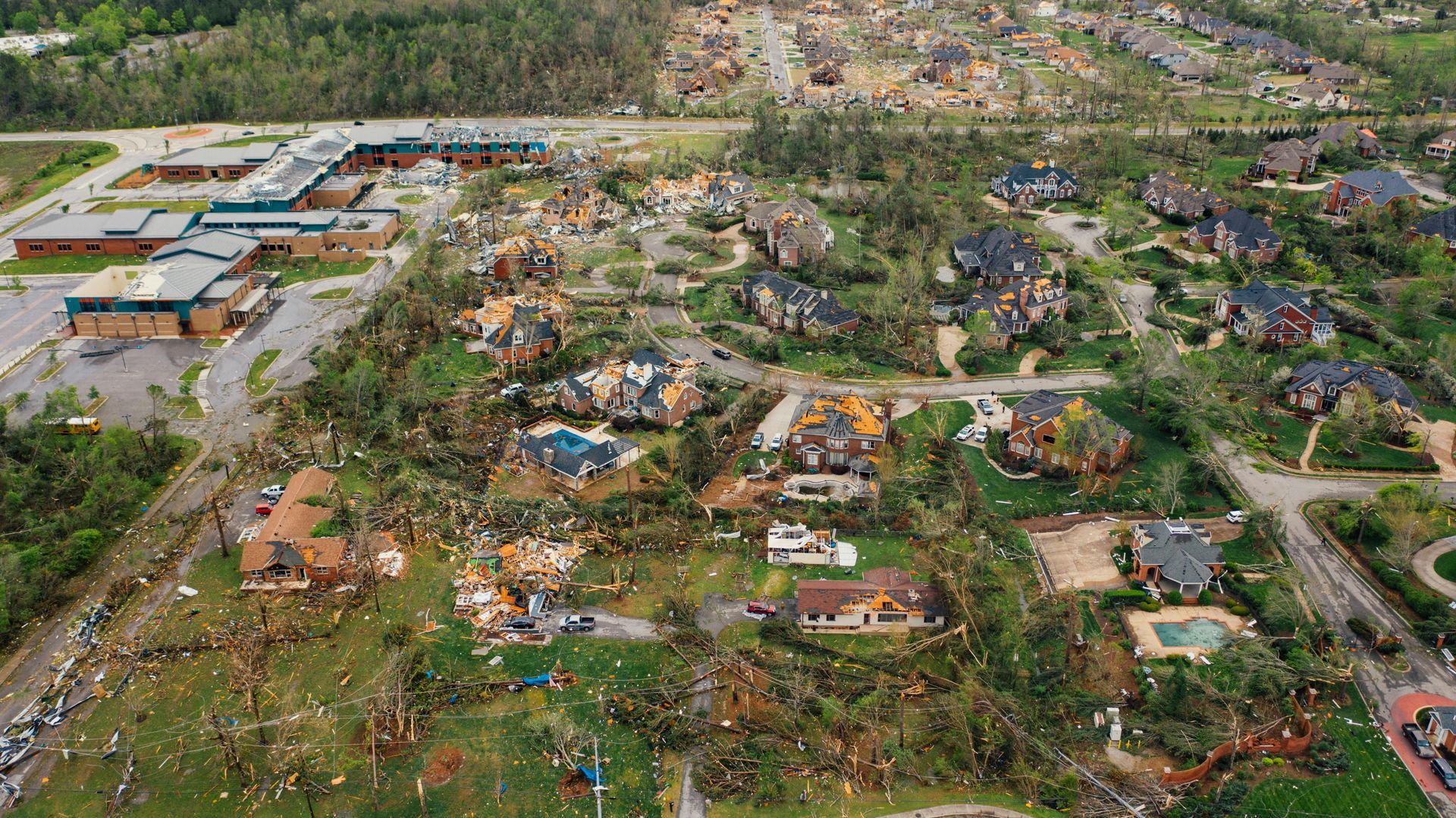 Aerial view of a neighborhood devastated by a tornado. Damaged homes and debris scattered across green lawns.