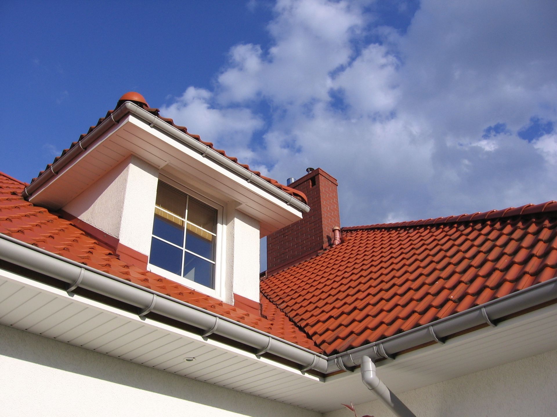 Red-tiled roof with a dormer window and chimney against a blue sky with fluffy white clouds.