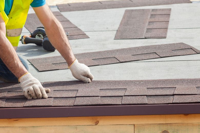 Roofer installing brown asphalt shingles on a roof, wearing gloves and a safety vest.