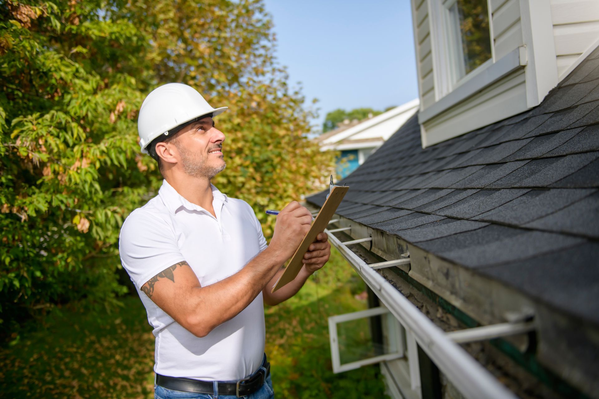 Man in hard hat inspecting a roof and gutter. He holds a clipboard, stands outdoors near a house.