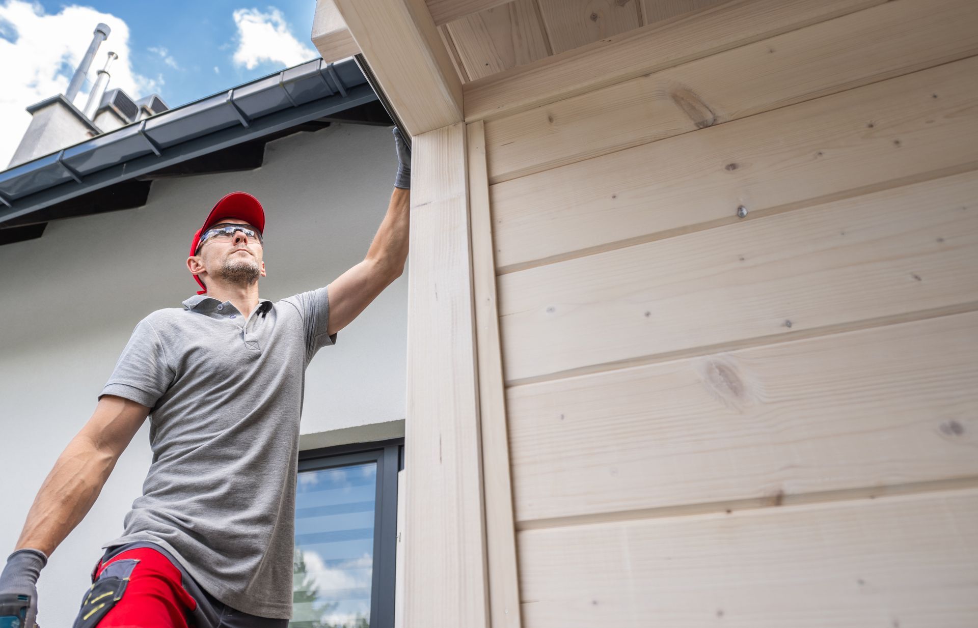 Man in red cap and gloves inspecting wooden siding on a house.
