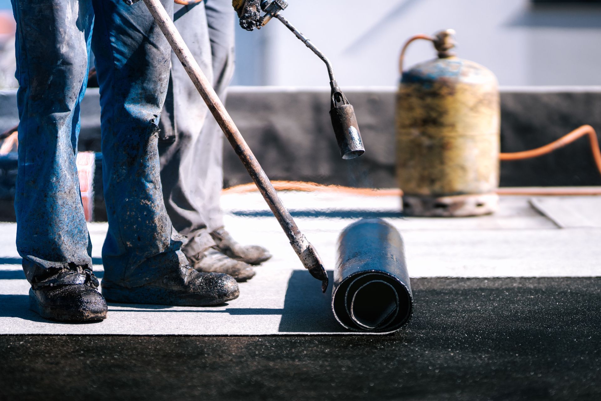 Two workers installing roofing with a torch and rolled material.