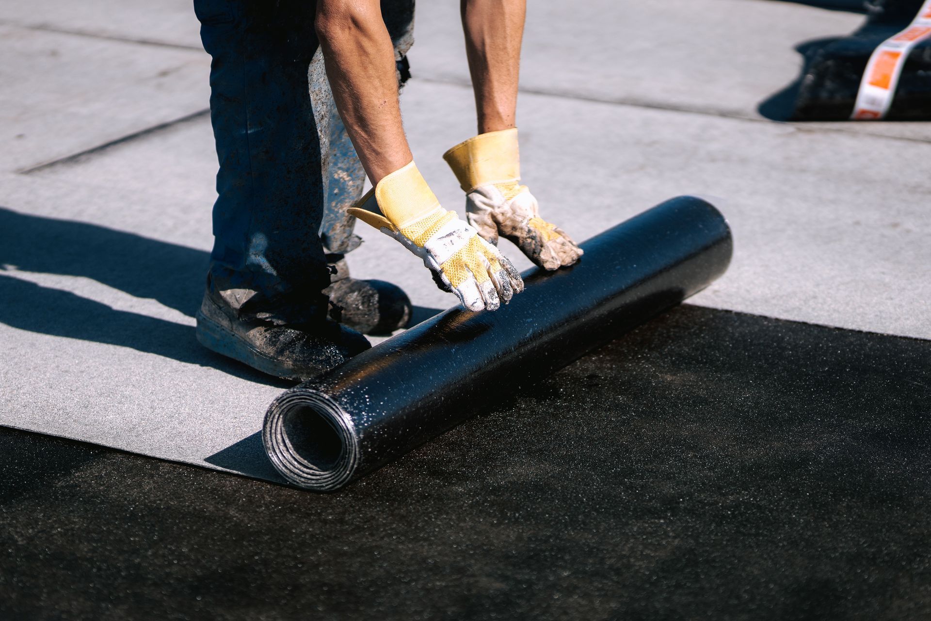 Person rolling out a black roofing material on a flat surface, wearing gloves.