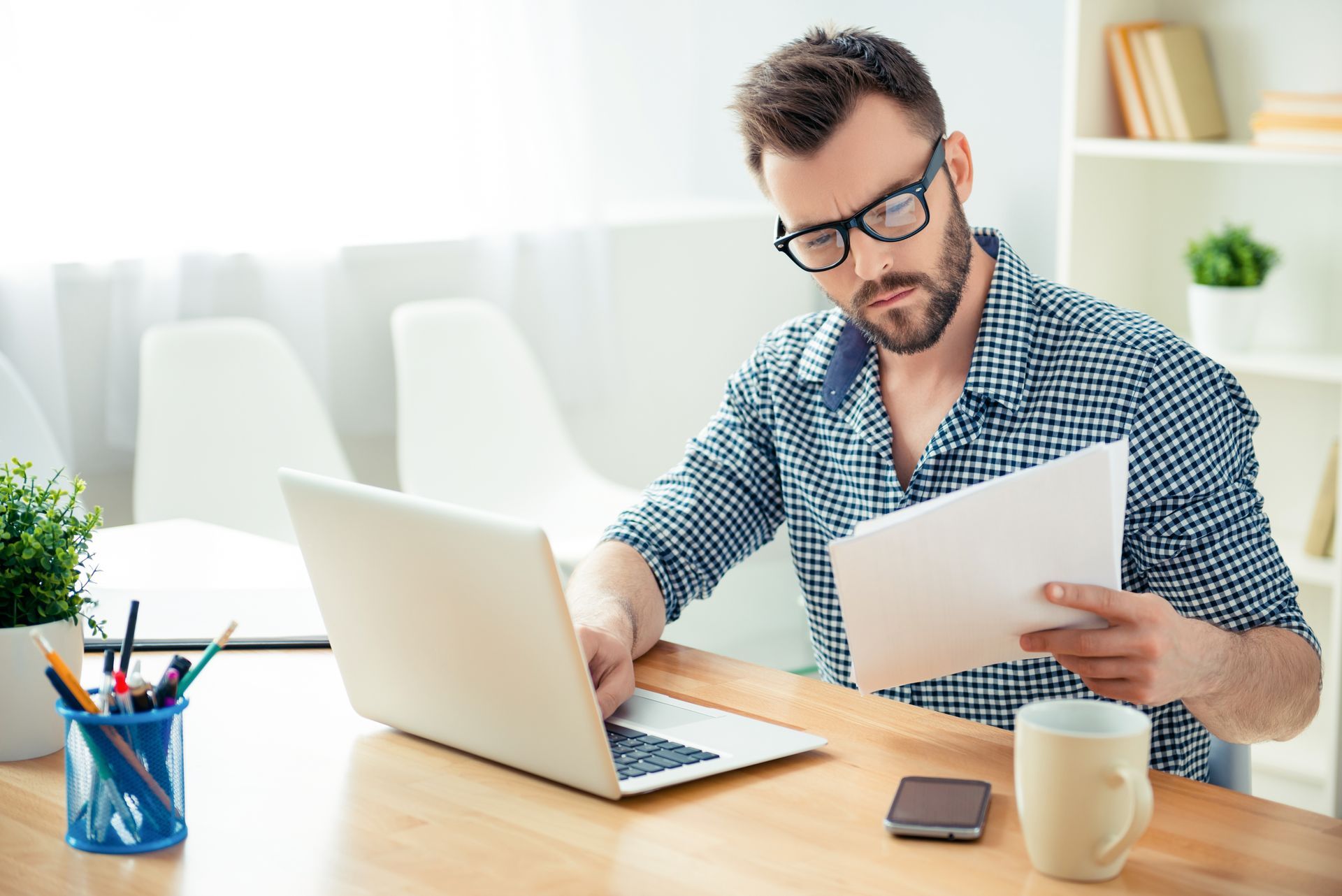 Man with glasses working on a laptop, holding papers, and looking at the screen. Wooden desk in a well-lit room.