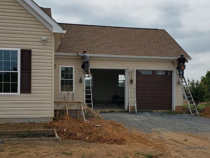 Two workers install gutters on a house under construction; beige siding, brown garage door, cloudy sky.