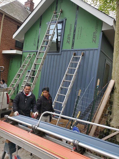 Two workers use a metal bender on siding. Blue siding and ladders are propped against a house under construction.