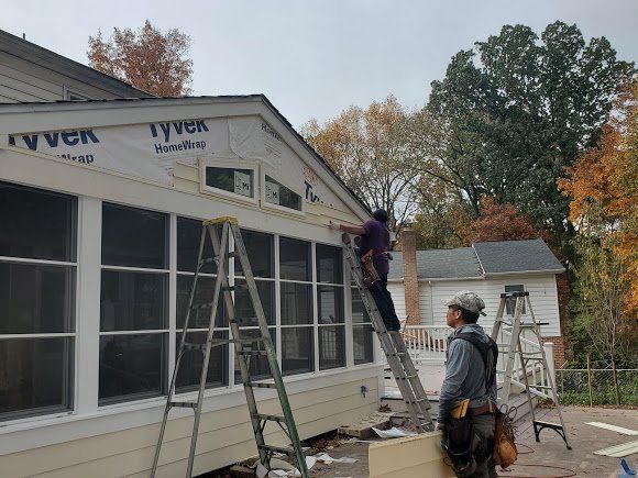 Two workers on ladders installing siding on a house. Overcast sky, fall foliage visible.