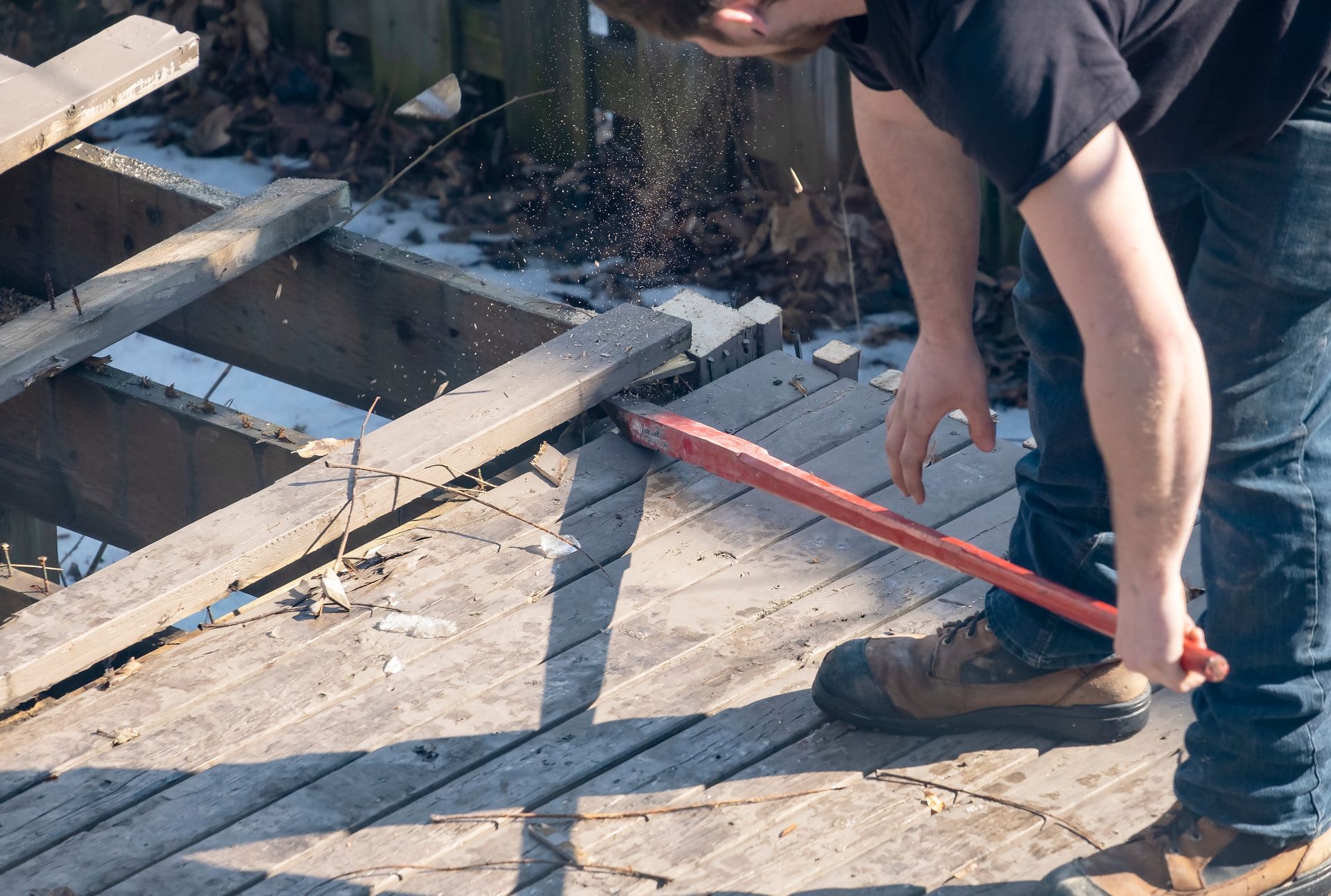 Person using a pry bar to remove wooden boards from a deck, outdoors.