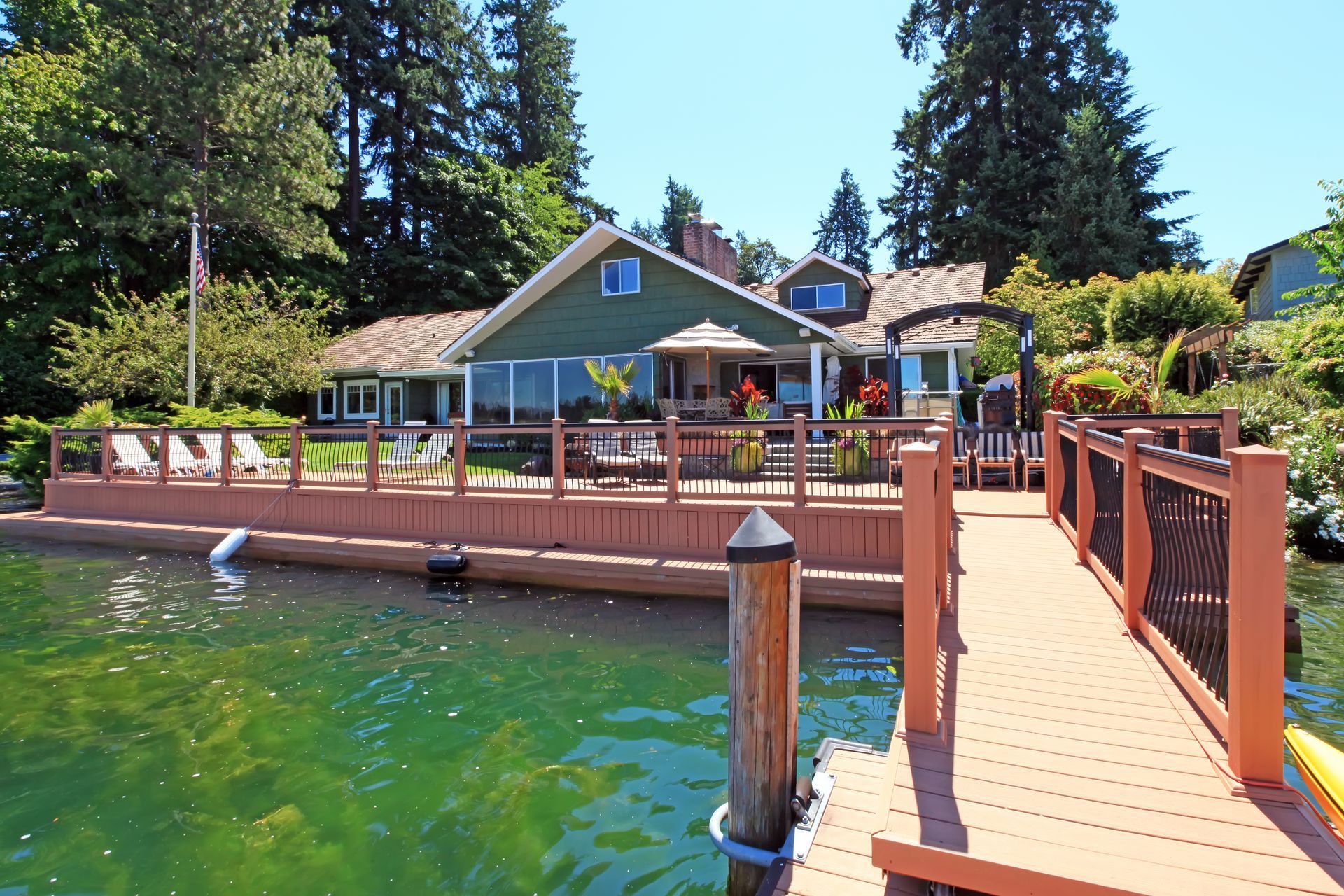 A green lake house with a wooden deck and dock. The water is green, with trees in the background.
