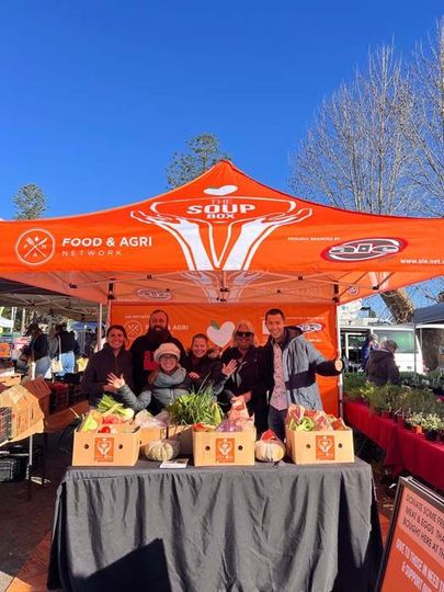 People at an orange FOOD & AGRI market tent displaying potted plants outdoors