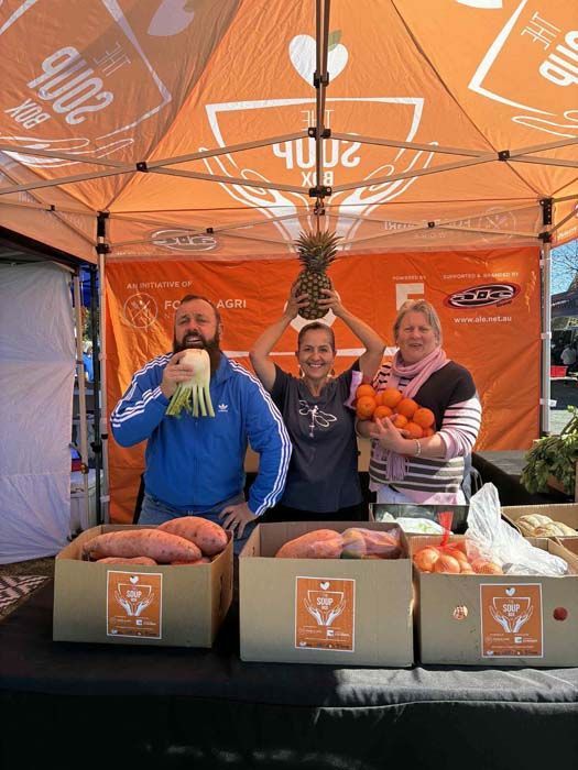 Three people under an orange market tent, standing behind boxes and produce on a table.