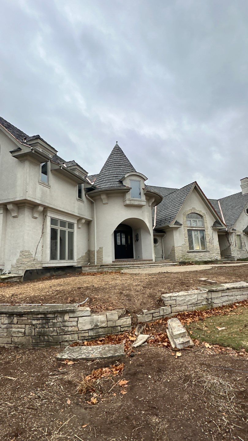 A large, light-colored French Provincial house with a turret entry and grey roof sits behind a low stone retaining wall.