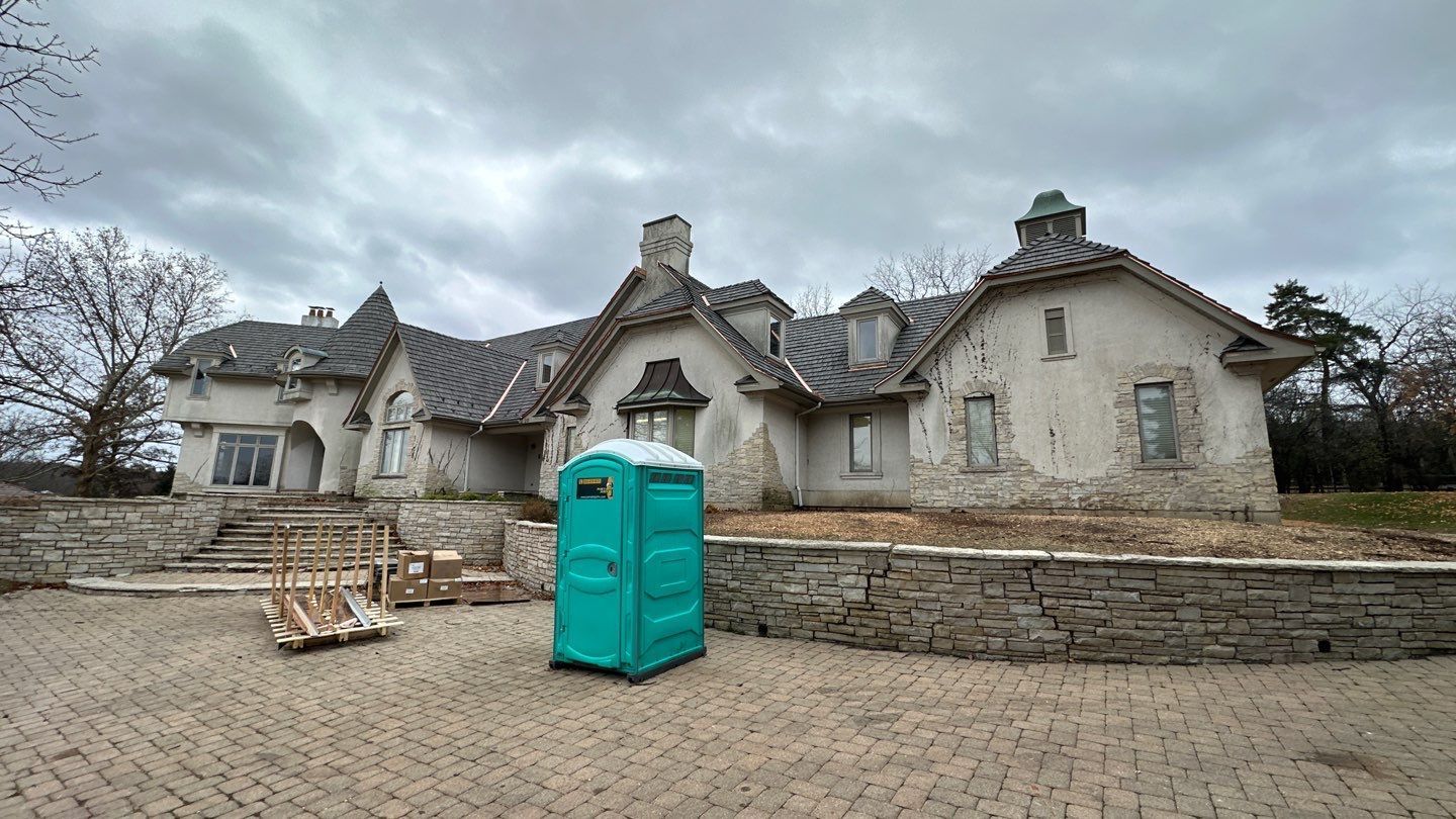 A bright green portable toilet stands on a paved patio in front of a large, weathered stone house under a cloudy sky.