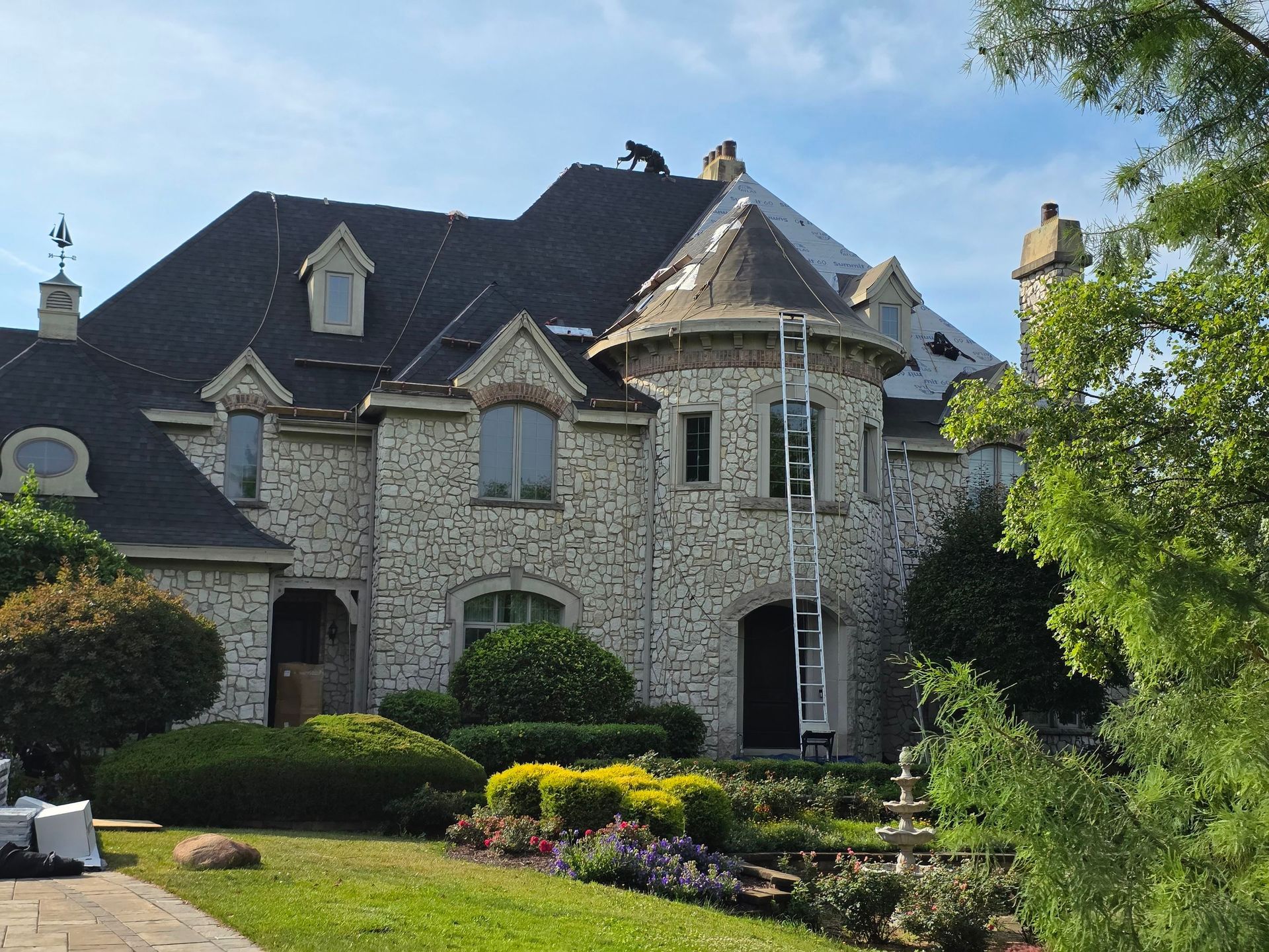 A stone-faced house with a circular turret tower, undergoing roof repairs with a ladder leaning against the exterior.