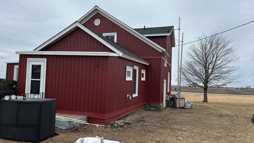 A red farmhouse with white trim sits on a grassy lot under an overcast sky, next to a bare tree and a tall antenna.