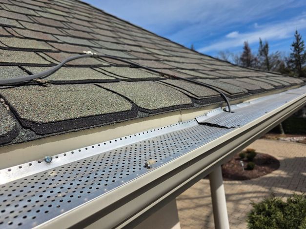 A perforated metal gutter guard installed on a sloped roof with shingles and a dark cable running across it.